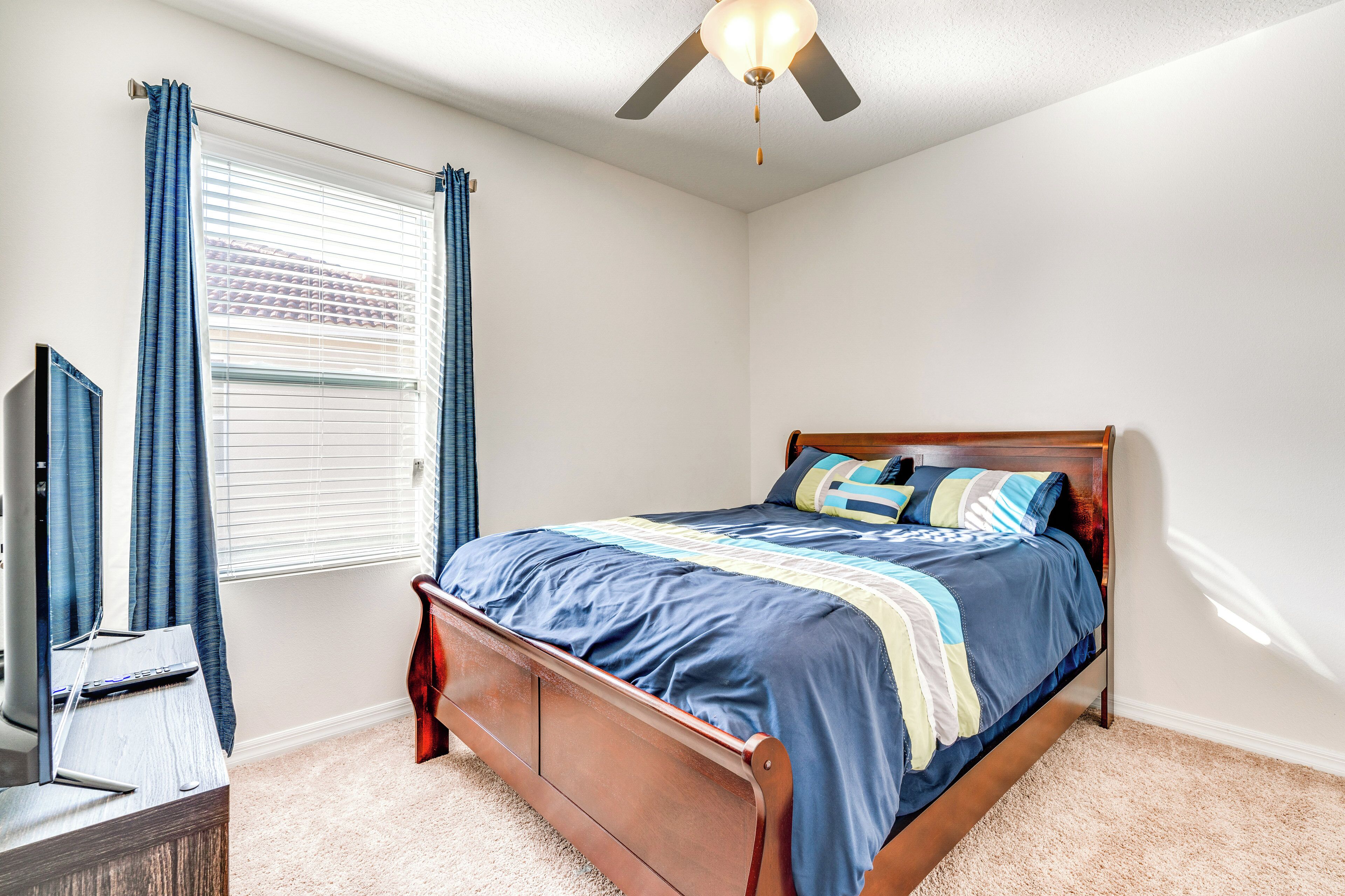 A bedroom with a wooden bed, a ceiling fan, a TV on a stand, and a window with curtains.