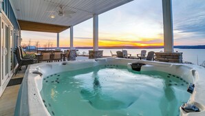 A hot tub with a view of the sunset over a body of water, surrounded by outdoor seating and a covered patio.