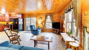 Interior acogedor de una cabaña con paredes y pisos de madera, un comedor, una sala de estar con sofá y sillones, una cocina con electrodomésticos y ventanas amplias.
