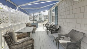 Een terras buiten met rieten meubilair, een wit hek en een overdekt gedeelte met een blauw zonnescherm.