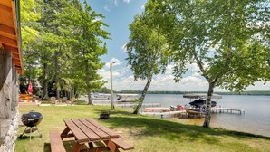 A lakeside picnic area with a wooden table and benches, surrounded by trees and a dock with boats. - South Twin Lake Cabin w/ Seasonal Dock & Slip! (Conover)