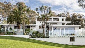 Una casa moderna de dos pisos con amplios ventanales y fachada blanca, rodeada de un césped cuidado y árboles.