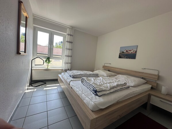 A bedroom with a bed, a window with curtains, a radiator, a picture on the wall, and a small table.