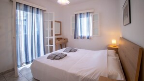 A neatly made bed with pillows and folded towels, a bedside table with a lamp, a wooden nightstand, and a window with curtains. - Naoussa Breeze Residence (Paros)