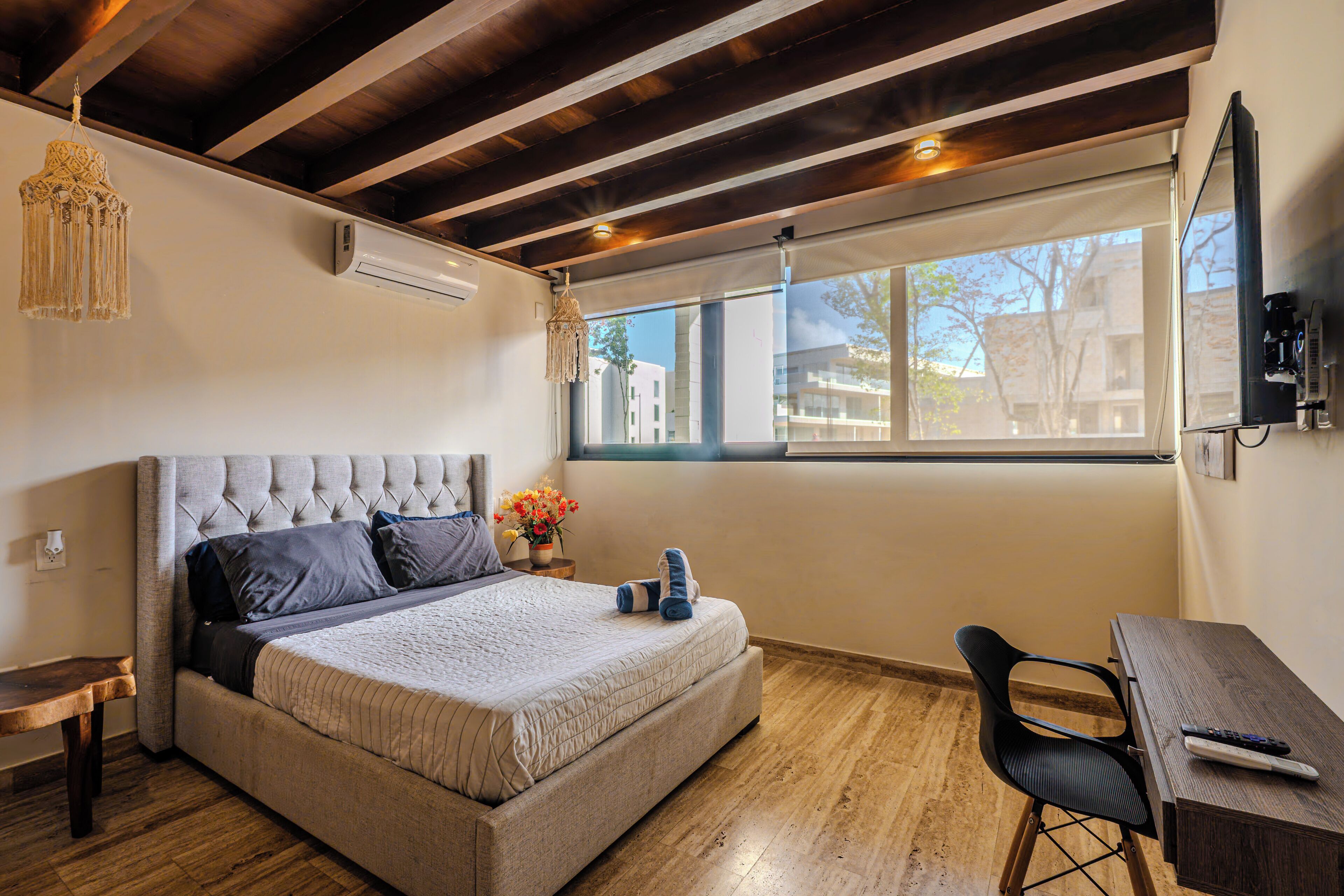 Un dormitorio con una cama grande, un escritorio de madera, una silla, una lámpara colgante y un ventanal con vistas a edificios.