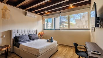 Un dormitorio con una cama grande, un escritorio de madera, una silla, una lámpara colgante y un ventanal con vistas a edificios.