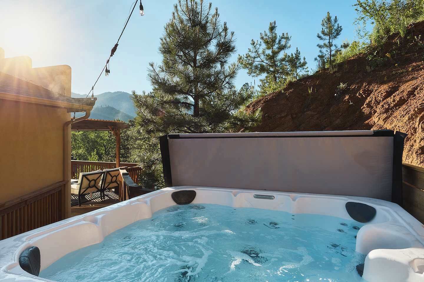 A hot tub with a wooden deck and chairs, surrounded by trees and a clear sky.