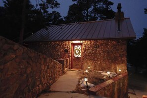Un bâtiment en pierre surmonté d’un toit en métal, une porte en bois avec un vitrage, et un chemin menant à l’entrée.