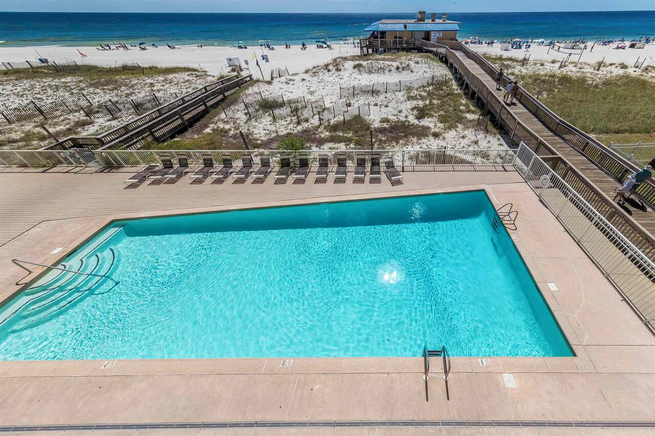 A rectangular pool with clear blue water, surrounded by a wooden deck and a wooden boardwalk leading to a beach with people and beach chairs.