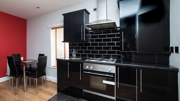 A modern kitchen with black cabinetry, a stainless steel oven, and a dining area with a red wall and black chairs.