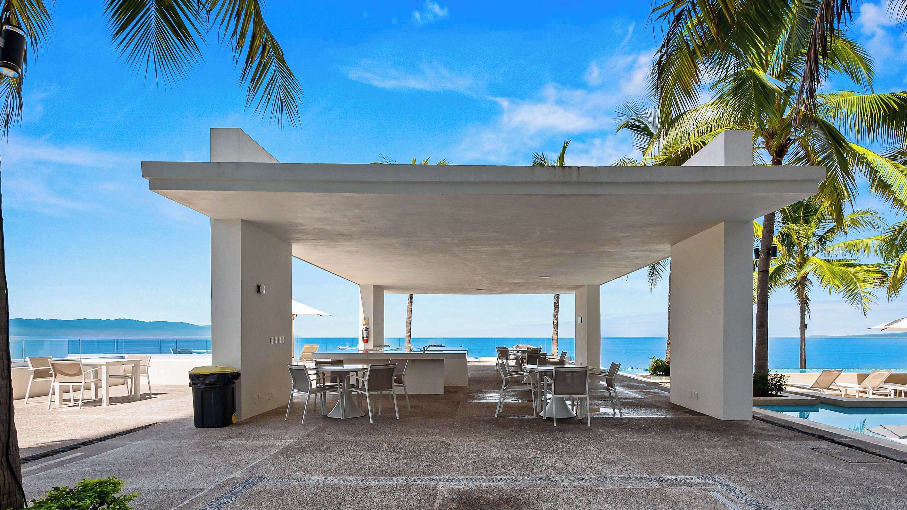 Ein überdachter Außenbereich mit Tischen und Stühlen, mit Blick auf einen Pool und das Meer.