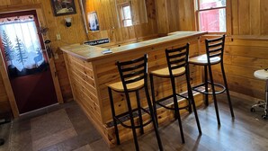 A wooden bar area with a sign reading "TEQUILLA TURNPIKE," three black bar stools, and a window with a view of trees. - Spacious Mio Cabin - Sleeps 20+ - Game Room - Near AuSable River & ORV Trails (Mio)