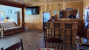 A cozy cabin interior with a sofa, a bar area with stools, a television mounted on the wall, and a clock. - Spacious Mio Cabin - Sleeps 20+ - Game Room - Near AuSable River & ORV Trails (Mio)