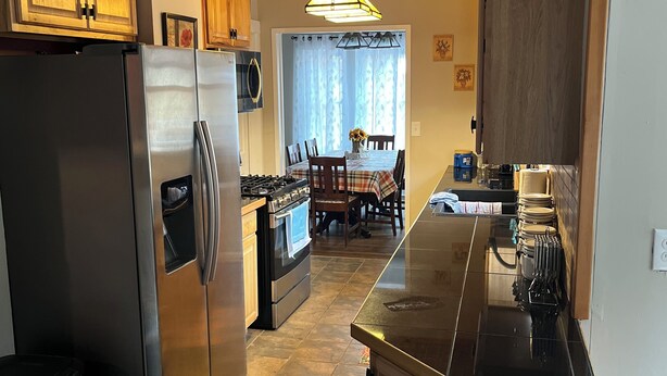 A kitchen with wooden cabinets, a stainless steel refrigerator, an oven, and a dining area with a table and chairs. - Spacious Mio Cabin - Sleeps 20+ - Game Room - Near AuSable River & ORV Trails (Mio)