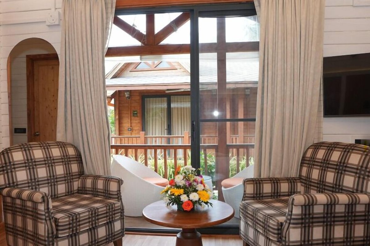Una habitación con dos sofás a cuadros, una mesa de madera con un centro de mesa floral y un ventanal con vistas a una terraza de madera y un edificio.