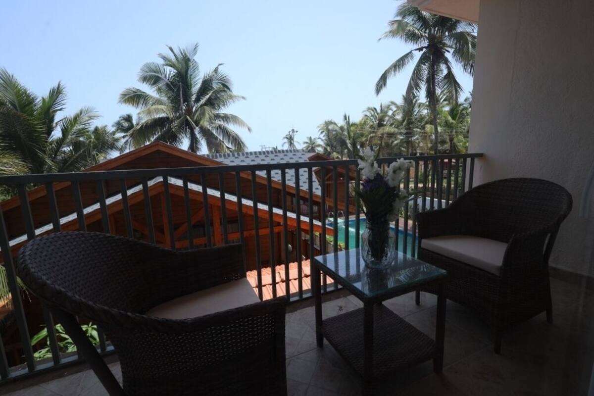 A balcony with wicker furniture and a glass table, overlooking a pool and palm trees.