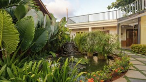 Un giardino con uno stagno, piante tropicali rigogliose e una scala in pietra che conduce a un balcone.