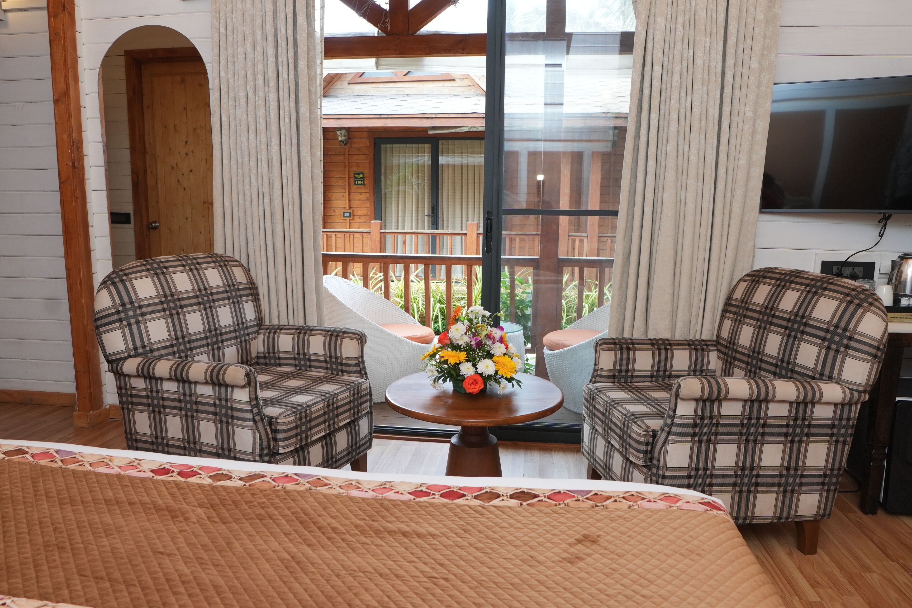 A room with two plaid armchairs, a small round table with a flower vase, and a bed with a quilted cover.