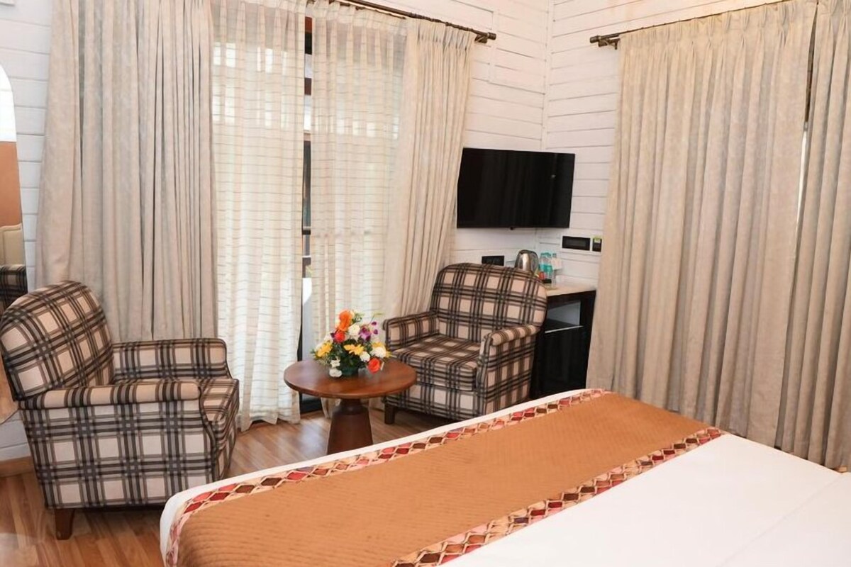 Habitación de hotel con cama, dos sillones a cuadros, una mesita con flores, televisor y cortinas.