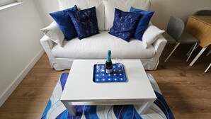 Ein weißes Sofa mit blauen Kissen mit Blumenmuster, ein weißer Couchtisch mit einer blauen Ablage, auf der sich eine Flasche und Gläser befinden, und ein grauer Sessel.