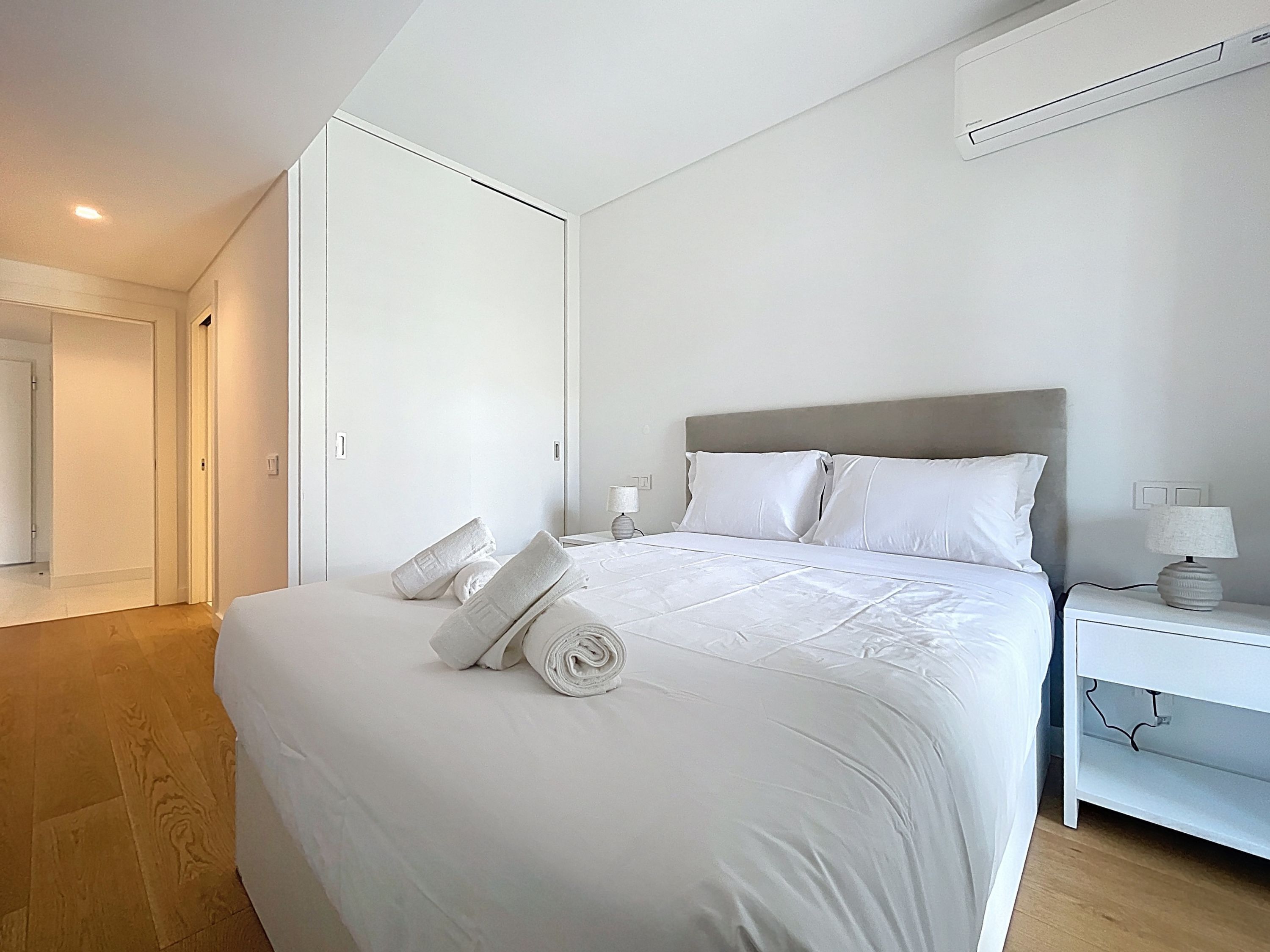 A modern hotel room with a neatly made bed, a bedside table with a lamp, and an air conditioning unit on the wall.