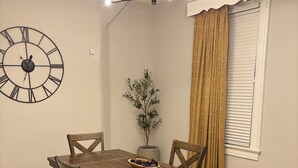 Een eetgedeelte met een houten tafel, vier stoelen, een wandklok, een plant en een raam met zonwering.