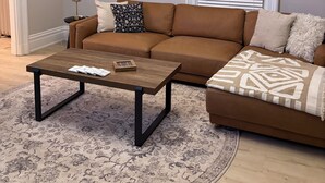 Een woonkamer met een bruine leren bank, een houten salontafel en een tapijt met dessin.