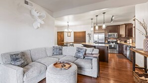 A modern living room with a sectional sofa, a dining area with a wooden table, and a kitchen with dark wood cabinets and stainless steel appliances. - Moose Mgmt at Bear Hollow Ridge 2207: Hot Tub, Pool, Hiking Trails, Shops-Dining (Park City)
