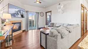 A modern living room with a fireplace, a sofa, a coffee table, and a large window with curtains. - Moose Mgmt at Bear Hollow Ridge 2207: Hot Tub, Pool, Hiking Trails, Shops-Dining (Park City)