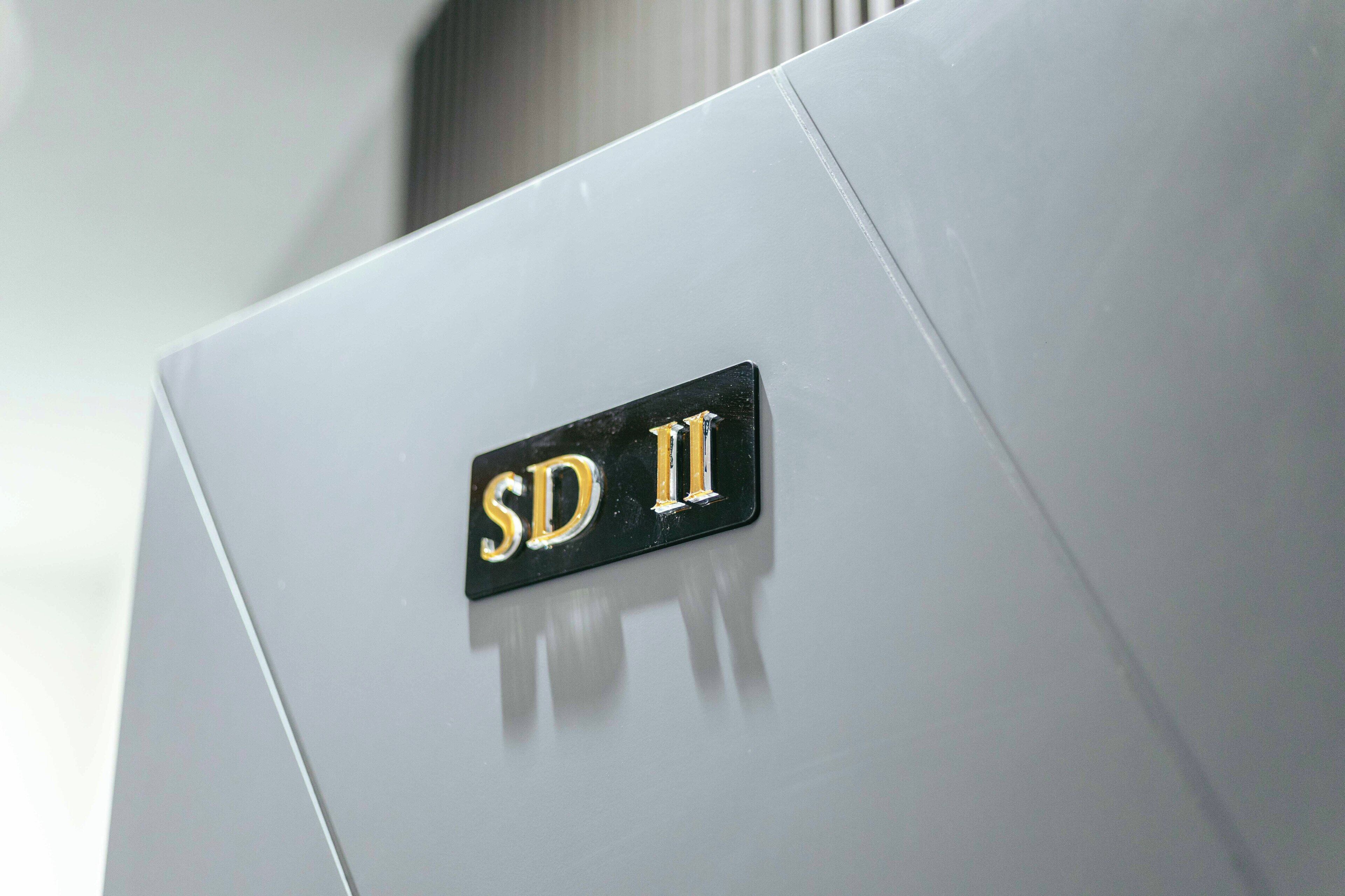 Een metalen badge met de tekst 'SD II' op een witte ondergrond.
