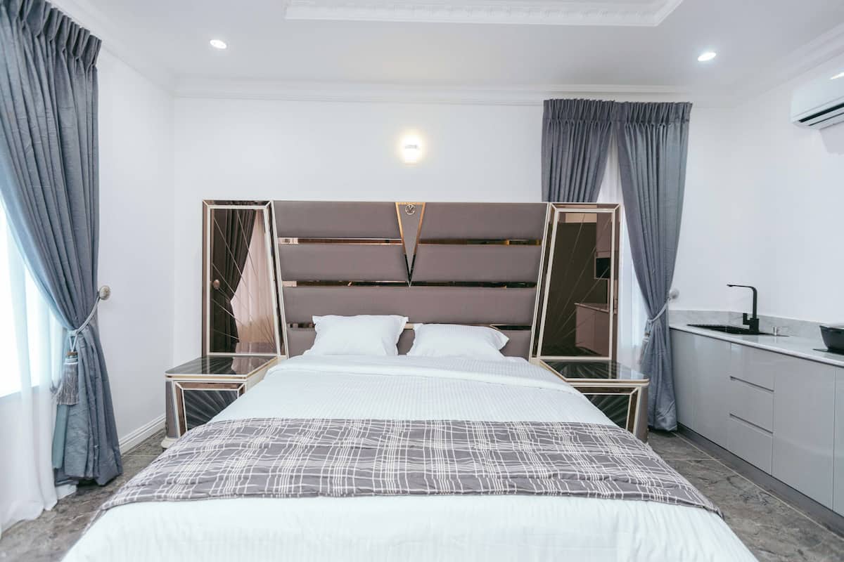 A modern hotel room with a large bed, a bedside table, a mirror, and a small kitchenette.