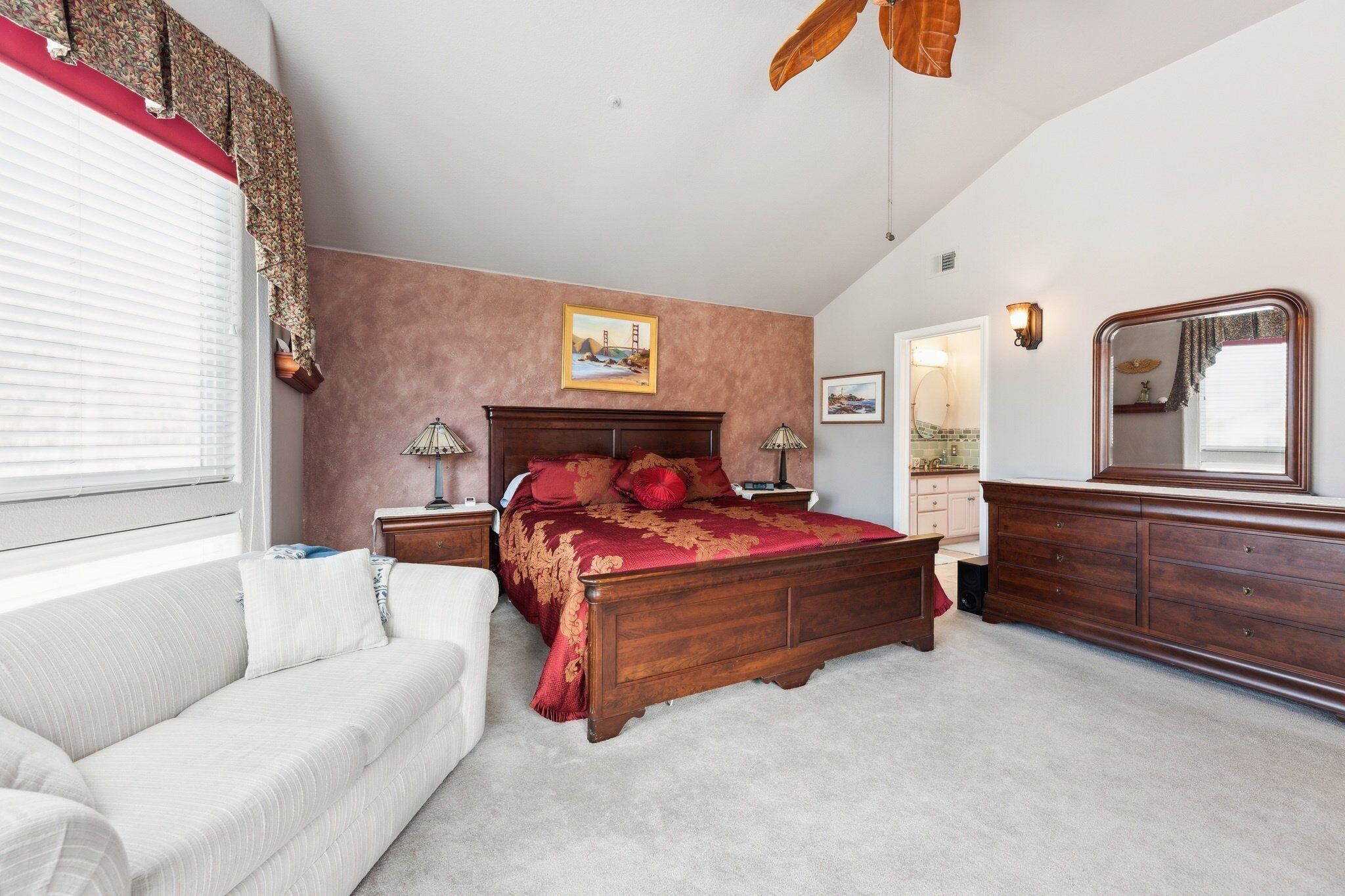 A bedroom with a large bed, a wooden dresser, a nightstand, a white sofa, and a ceiling fan.