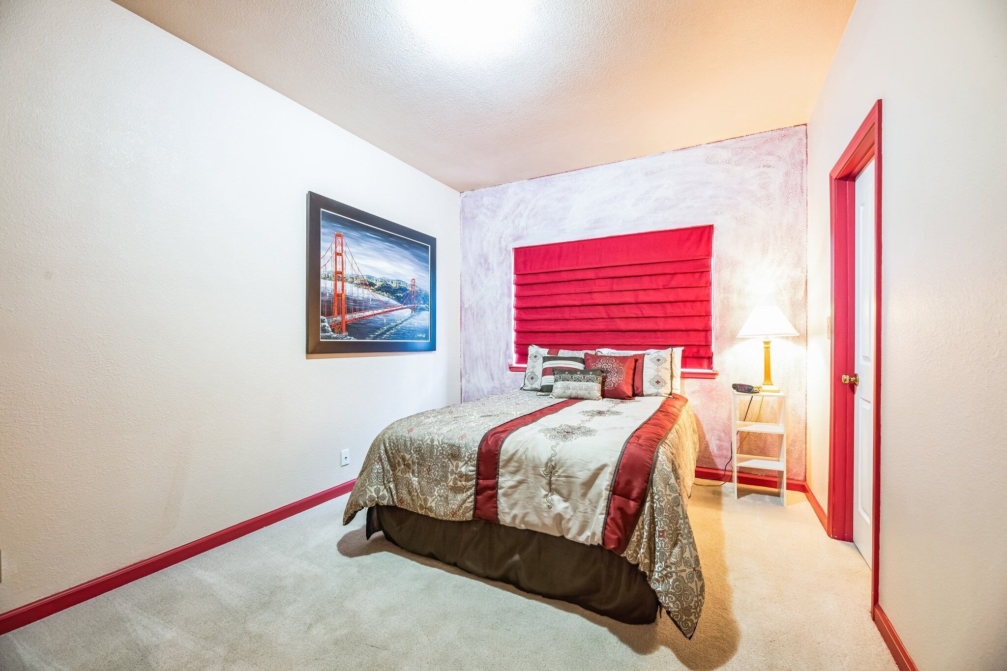 A bedroom with a bed, a red headboard, a nightstand with a lamp, a framed picture on the wall, and a red door.