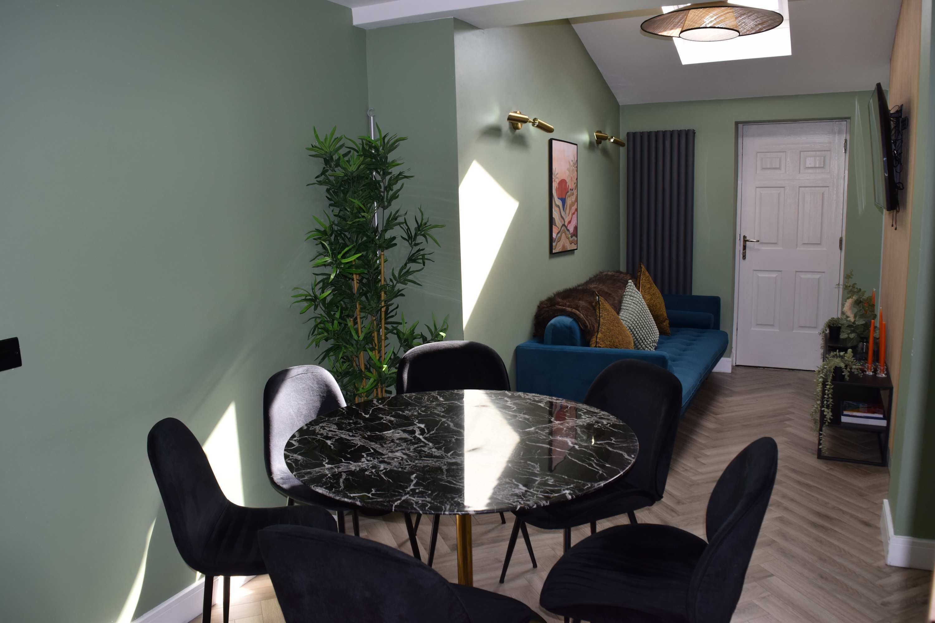 A dining area with a round table and chairs, a sofa, a potted plant, and a door in the background.
