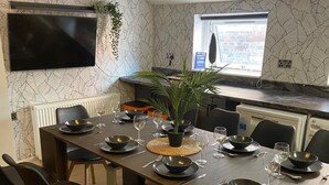 Un espace repas moderne avec une longue table dressée pour six personnes, entourée de chaises noires. Un téléviseur à écran plat est fixé au mur, au-dessus de la table.