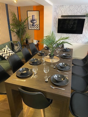 A dining area with a wooden table set for six, surrounded by black chairs. There are potted plants on the table, and a television mounted on the wall above.
