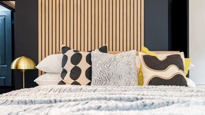 Un lit recouvert d’une couverture blanche à texture, une lampe de sol dorée et une variété d’oreillers à motifs.