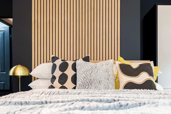 A bed with a white textured blanket, a gold floor lamp, and a variety of patterned pillows.