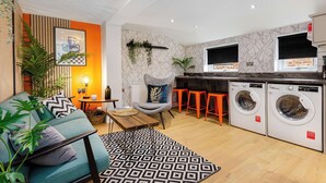 Un espace de vie compact comprenant un canapé, une table basse, un fauteuil, un lave-linge et un coin cuisine.