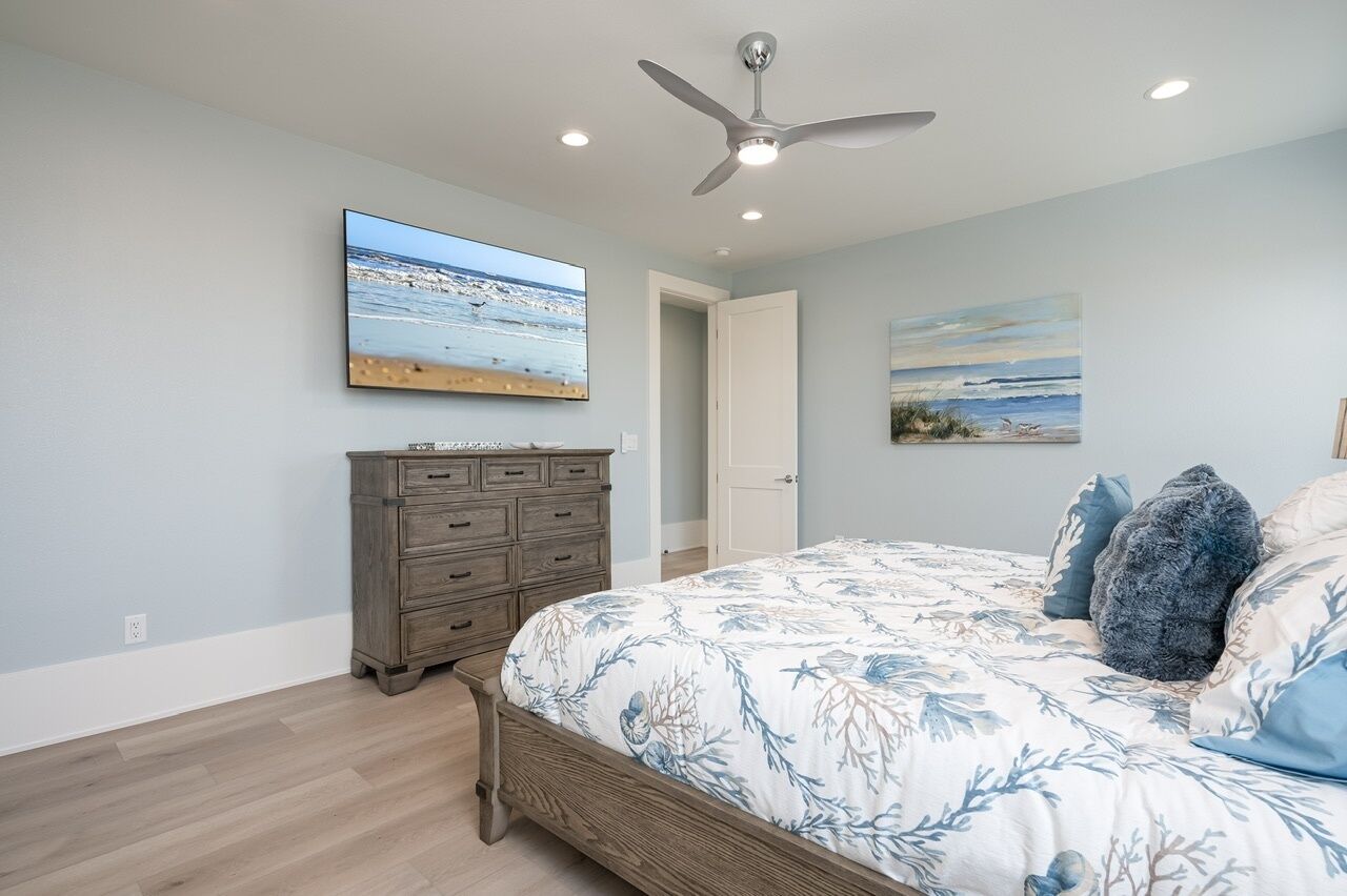 A bedroom with a bed, a dresser, a ceiling fan, and two paintings on the walls.