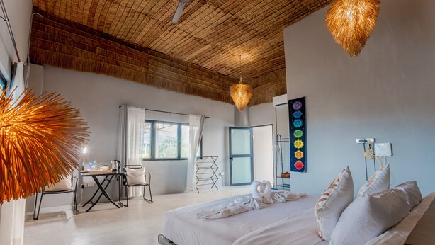 Une chambre d’hôtel avec un lit, un bureau et une chaise. On y trouve un ventilateur de plafond et un luminaire fixé au mur.