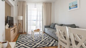 A living room with a grey sofa, a white chair, a TV on a wooden stand, a lamp, and a patterned rug. - Seaview Retreat for 6 people (Tkon)
