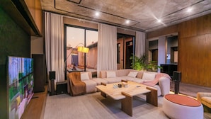 A modern living room with a sectional sofa, a coffee table, and a round ottoman. There is a television on the left and a plant in the corner. - Portal Chalets With Private Pool-Al Arid (Riyadh)