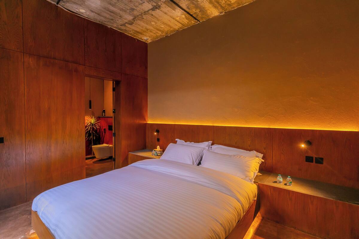 Ein Schlafzimmer mit einem großen Bett, Holzvertäfelung, einem Nachttisch mit Lampe und einem kleinen Tisch mit Flaschen.