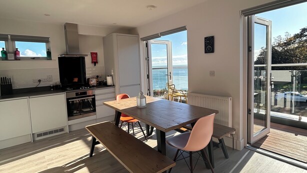 A modern kitchen with a dining area, a large window with a view of the sea, and a sliding glass door to an outdoor balcony. - The Beach Retreat Apartment, Balcony views of shanklin bay. (Shanklin)