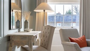 Una habitación con un escritorio de madera, una silla, un sillón a rayas, una lámpara y un ventanal con cortinas.