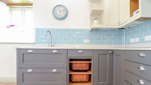 Una cocina con gabinetes grises, salpicadero de azulejos azules, un reloj de pared y cestas de mimbre en un mueble.