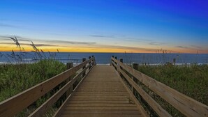 A wooden boardwalk leading to a beach with a clear view of the ocean and a sunset in the background.