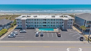 Un hotel con piscina en la azotea, un estacionamiento con varios coches y vistas a la playa y al océano.