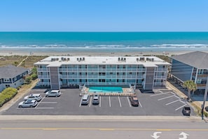 A hotel with a rooftop pool, a parking lot with several cars, and a view of the beach and ocean.